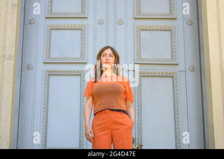 Low-Angle-Ansicht einer Frau in farbenprächtigem orangefarbenem Outfit, die mit Händen in den Taschen vor einer großen historischen Holztür mit Kopffarbe wartet Stockfoto