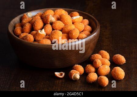 Paprika beschichtete Erdnüsse in einer braunen Holzschale neben verschütteten Erdnüssen auf dunklem Holz. Stockfoto