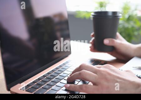 Weibliche Hände, die auf der Laptop-Tastatur tippen, halten Glas mit Kaffee Stockfoto