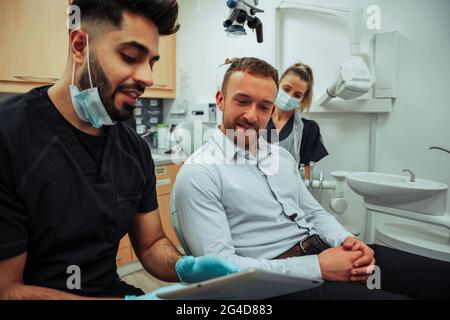 Kaukasischer männlicher Geschäftsmann, der die Arztpraxis für die jährliche Untersuchung besucht. Arzt und Ärztin, die im Klinikraum auf einem digitalen Tablet scrollt Stockfoto