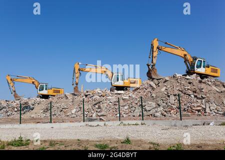 Drei gelbe Bagger auf einem Schmutzhaufen hinter dem Zaun, mit blauem Himmel im Hintergrund Stockfoto