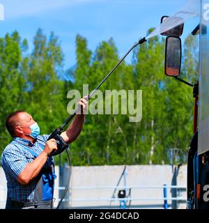 Autowaschanlage für Sonderausrüstung und LKW. Ein männlicher Fahrer mit einer medizinischen Maske wäscht an einem Sommertag unter Druck auf der Straße einen Lastwagen mit einer Pistole. Stockfoto