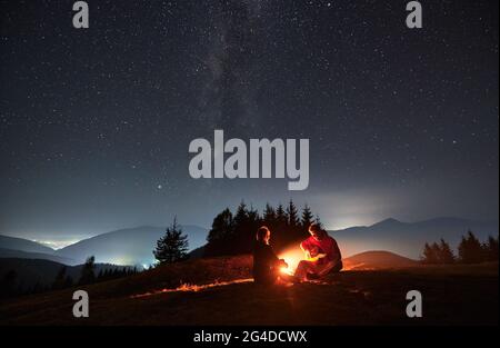 Junger Mann, der am Lagerfeuer auf dem Berg sitzt und Gitarre für seine Geliebte unter dem Sternenhimmel der Nacht mit der Milchstraße spielt. Silhouetten von Bäumen, Bergen und Licht aus umliegenden Dörfern im Hintergrund Stockfoto