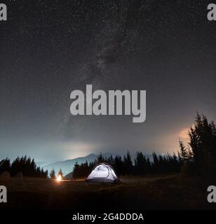 Helles Zelt in der Nähe von Wald und Lagerfeuer. Gemütlicher Platz für Camping in den Bergen unter strahlendem Himmel mit Sternen und Milchstraße bedeckt. Silhouetten von Berggipfeln im Hintergrund. Stockfoto