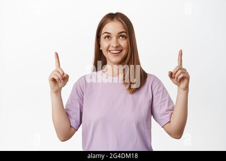 Portrait eines glücklichen blonden Mädchens, das nach oben zeigt, lächelt und auf den Werbetext des Logos schaut, das Schild oben liest und im T-Shirt gegen Weiß steht Stockfoto