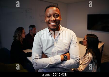 Porträt eines lächelnden afrikanischen Geschäftsführers, der vor seinem Kollegen posiert, der im Hintergrund arbeitet Stockfoto