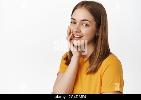 Porträt eines schönen Teenagers, Haut ohne Akne berühren, kein Make-up, Wange sanft berühren und zart lächelnd vor der Kamera, im gelben T-Shirt stehend Stockfoto