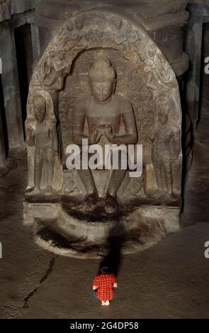 Ein Mädchen in einem roten Kleid sitzt zu den Füßen des Buddha und betet. Es gibt eine sehr große und lineare Statue von Buddha, Ellora Höhlen, Aurangabad-in Stockfoto