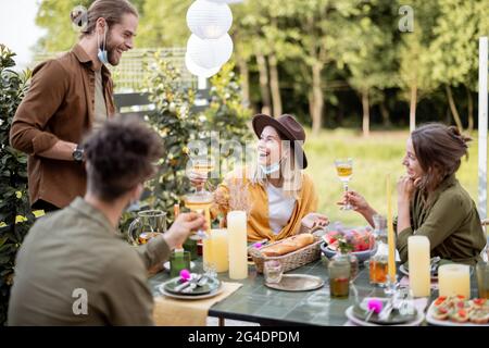 Junge Freunde in Gesichtsmaske bei einem festlichen Abendessen während eines Sommers im Hinterhof des Landhauses. Glücklich, sich im wirklichen Leben danach zu treffen Stockfoto