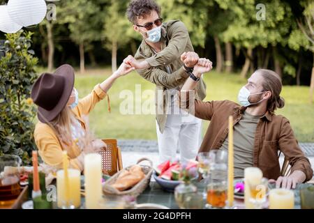 Eine Gruppe von Freunden, die während eines Mittagessens im Freien eine Gesichtsmaske tragen, sich gegenseitig begrüßen und dabei soziale Distanz bewahren, während einer Pandemie Zeit miteinander verbringen Stockfoto