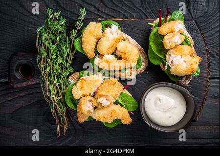Toasts mit knusprig gebratenen Garnelen und grünem Salat auf einem Holzbrett. Schwarzer Holzhintergrund. Draufsicht Stockfoto