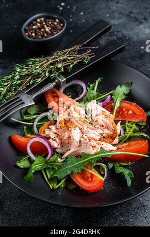 Salat mit gebackenem Lachsfiletsteak, frischer Rucola und Tomate auf einem Teller. Schwarzer Hintergrund. Draufsicht Stockfoto