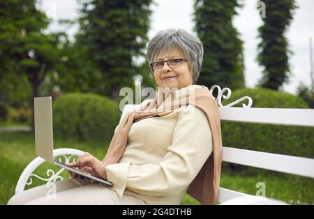 Porträt einer glücklichen älteren Dame, die auf einer Parkbank sitzt und ihren Laptop benutzt Stockfoto