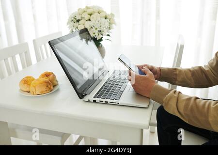 Hände eines reifen Mannes, der Nachrichten auf dem Smartphone beantwortet, wenn er zu Hause am Küchentisch arbeitet, weil er eine Coronavirus-Pandemie hatte Stockfoto
