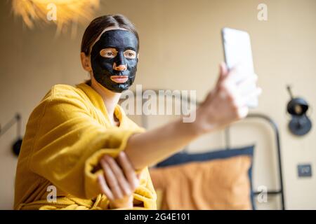 Frau im Bademantel mit schwarzer Reinigungsmaske und Telefon im Schlafzimmer Stockfoto