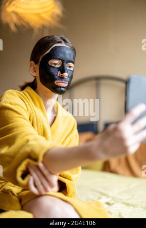 Frau im Bademantel mit schwarzer Reinigungsmaske und Telefon im Schlafzimmer Stockfoto