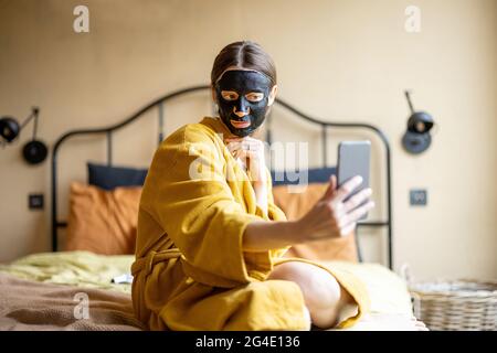 Frau im Bademantel mit schwarzer Reinigungsmaske und Telefon im Schlafzimmer Stockfoto
