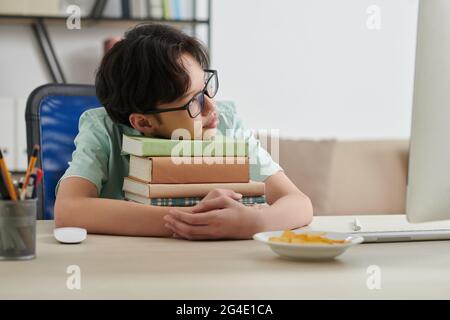 Träumende Teenager-Schuljunge in Brille auf Stapel von Studenten Bücher auf seinem Schreibtisch zu Hause gelehnt Stockfoto