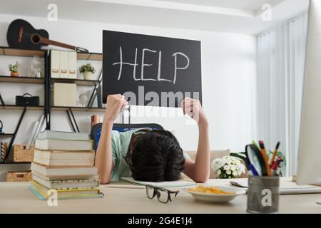 Erschöpfter Schuljunge, der es leid ist, am Schreibtisch zu sitzen und ein Hilfsschild zu zeigen Stockfoto
