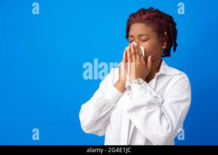 Kranke Afro-Frau niest in Papierserviette vor blauem Hintergrund Stockfoto