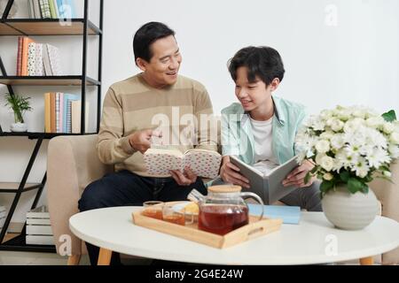 Fröhlicher Vater und Teenager-Sohn lesen und diskutieren Bücher mit interessanten Romanen zu Hause beim Tee trinken Stockfoto