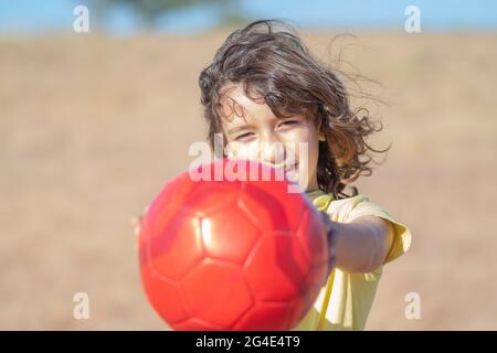 Kleiner Junge mit langen Haaren und gelbem T-Shirt spielt an einem sonnigen Sommertag mit seiner roten Kugel auf einem Feld Stockfoto