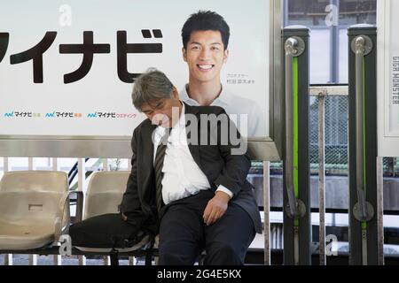 Ein Geschäftsmann, der auf einem Bahnsteig schläft, vor einem Plakat eines lächelnden jungen Mannes in Tokio, Japan Stockfoto