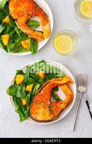 Gebratenes Lachssteak mit Spinat und gelben Tomaten auf einem Teller auf grauem Grund, ein Glas Wasser mit Zitrone, ein herzhaftes Mittagessen mit gesundem rotem Fisch Stockfoto