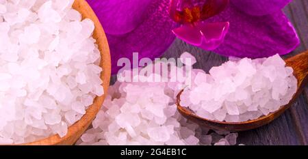 Weißes Badesalz in einem Holzbecher mit einem Löffel auf einem Holztisch mit einer Orchidee. Stockfoto