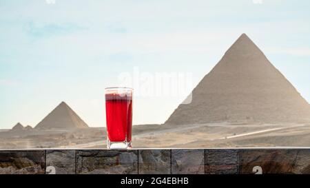 Traditionelle arabische Kräutertee-Karkade im Glas vor dem Hintergrund der Großen Pyramiden von Chephren Khufra und Cheops Khufu in Gizeh. Wunderschöne Aussicht auf Stockfoto