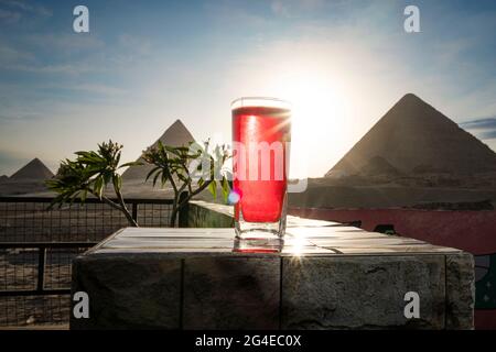 Traditionelle arabische Kräutertee-Karkade in einem Glas vor dem Hintergrund der Großen Pyramiden von Gizeh. Schöne Aussicht auf die Attraktion im Restaurant Stockfoto