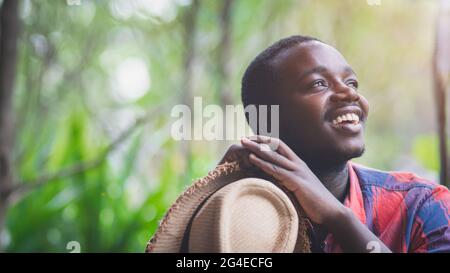 Glücklicher afrikanischer Mann lächelt und hält Hut mit dem grünen Natur Hintergrund Stockfoto