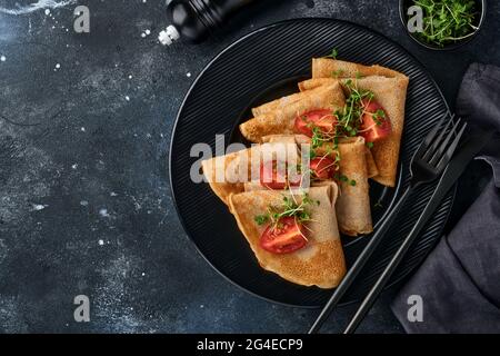 Stapel von glutenfreien Buchweizenmehl Crepes Pfannkuchen mit Kirschtomaten und Rucola Microgreens auf schwarzem Teller auf dunklem Hintergrund, hausgemacht gesund Stockfoto