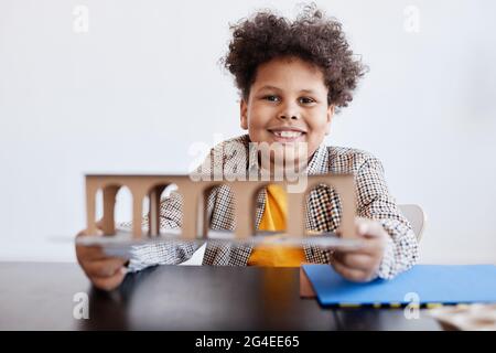 Vorderansicht Porträt eines lächelnden afroamerikanischen Jungen, der ein Pappmodell hält, während er an einem handgefertigten Schulprojekt arbeitet, Kopierraum Stockfoto
