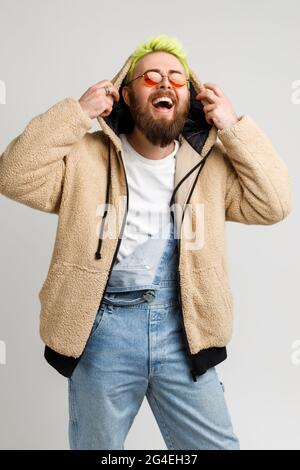 Extrem glücklicher bärtiger junger Erwachsener, der Denim-Overalls und -Jacke trägt, laut lacht, die Hände auf seiner Kapuze hält, hört lustigen Witz. Stehbolzen für den Innenbereich Stockfoto