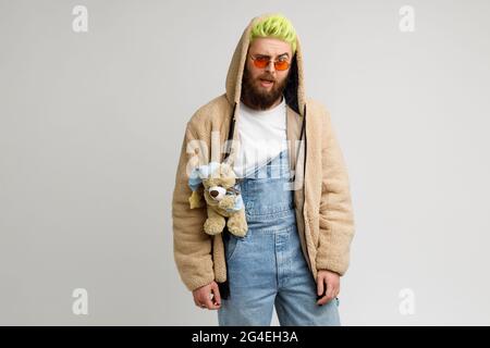 Mann mit Bart Model trägt trendige Denim Overalls und Jacke, Blick auf die Kamera mit rätselhaften Gesichtsausdruck, angehoben Augenbraue. Studio im Innenbereich Stockfoto