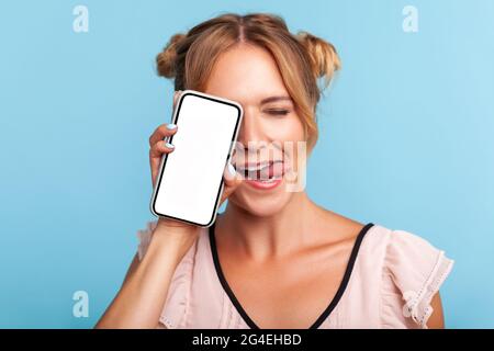 Porträt einer schönen positiven blonden Frau mit zwei lustigen Haarbrötchen im Kleid stehend, die das halbe Gesicht mit dem Telefon bedeckt und die Augen schließt und die Zunge zeigt. Zoll Stockfoto