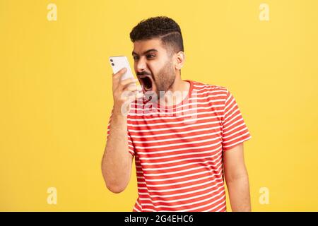 Wütender, verrückter Mann mit Bart im gestreiften T-Shirt, laut schreiendes Telefon, was die Sprachnachricht mit wütendem, aggressivem Ausdruck hinterlässt. Studio-Zimmer im Innenbereich Stockfoto