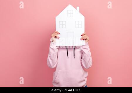 Porträt einer jungen Frau im Kapuzenpullover-Gesicht mit großem Papierhaus, träumt von einer eigenen Unterkunft. Innenaufnahme des Studios isoliert auf rosa Hintergrund Stockfoto