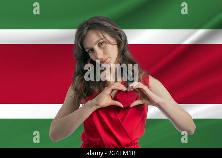 Ich Liebe Suriname. Ein Mädchen hält ein Herz auf ihrer Brust in ihren Händen vor dem Hintergrund der Flagge von Suriname. Der Begriff des Patriotismus. Stockfoto