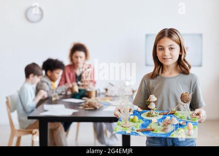 Waist-up-Porträt eines lächelnden Schulmädchen mit Model und Blick auf Kamera während Kunst- und Handwerksunterricht in der Schule, kopieren Raum Stockfoto