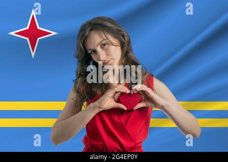 Ich Liebe Aruba. Ein Mädchen hält ein Herz auf ihrer Brust in ihren Händen vor dem Hintergrund der Flagge von Aruba. Der Begriff des Patriotismus. Stockfoto