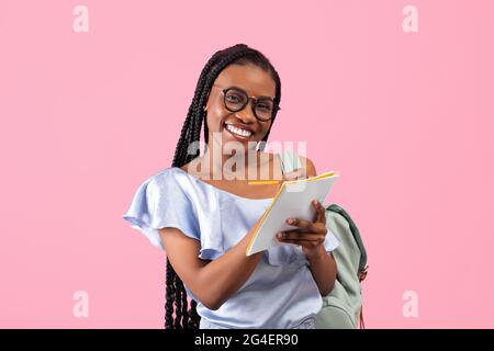 Smart junge schwarze Frau mit Rucksack tragen Brille, Notizen über rosa Studio-Hintergrund. Hochschulbildung Stockfoto