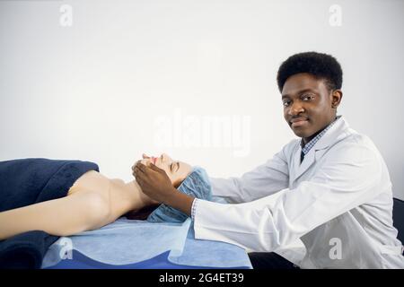 Hübscher junger professioneller afroamerikanischer Mann Masseur des medizinischen Zentrums, der eine Gesichtsmassage mit Anti-Aging-Behandlung für junge hübsche weibliche Klientin durchführt, die auf dem Massageliegen liegt Stockfoto