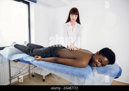 Junge Ärztin Physiotherapeutin massiert einen jungen afro-amerikanischen Mann auf den Rücken in der Arztpraxis. Entspannender junger Mann, der auf der Couch liegt und eine Massagetherapie genießt. Stockfoto