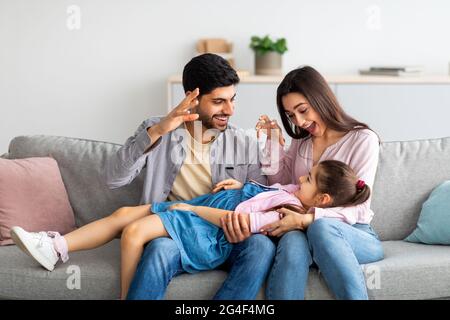 Fröhliche junge östliche Eltern verbringen Zeit mit ihrer kleinen Tochter zu Hause und spielen mit Kindergunst auf der Couch Stockfoto