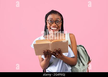 Aufgeregt junge afroamerikanische Studentin mit Rucksack, der offenes Buch auf rosa Hintergrund hält Stockfoto