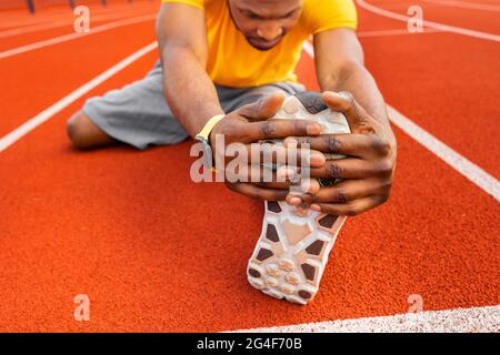 Nahaufnahme eines jungen afroamerikanischen Mannes, der am Stadion sitzt und sich auf hellem Hintergrund die Beine streckt. Professioneller junger schwarzer Mann in gelber Sportkleidung Stockfoto