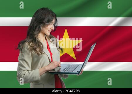 Freiberuflich in Suriname. Schöne junge Frau Freiberuflerin nutzt Laptop-Computer vor dem Hintergrund der Flagge von Suriname. Stockfoto