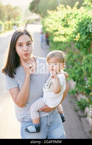 Mama macht Gesichter und hält ein kleines Mädchen in ihren Armen am Zaun, umschlungen von Efeu Stockfoto